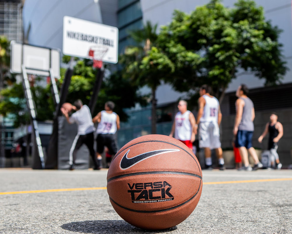 A Nike-branded basketball lies on the asphalt, while a Nike 3-on-3 game takes place out of focus in the background.